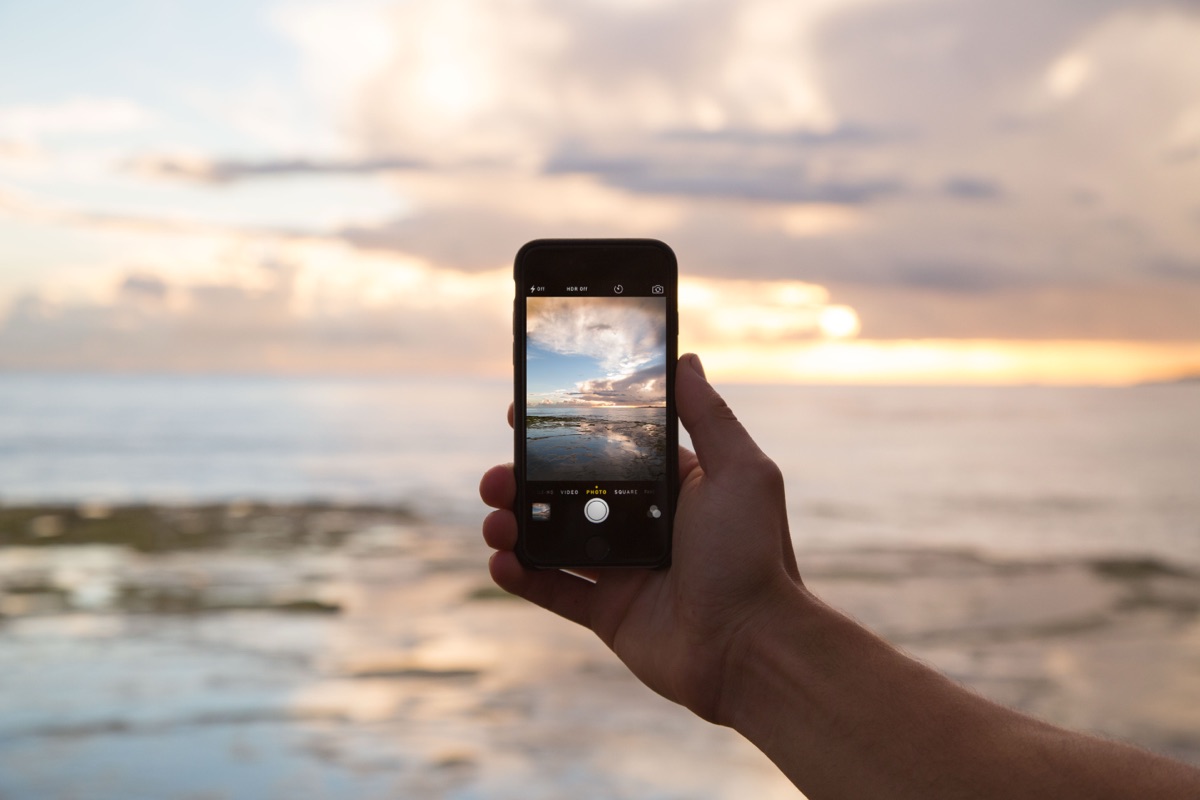 A hand holding an iPhone up to photograph a glowing sunset sky, illustrating the 2026 iPhone Photography Awards