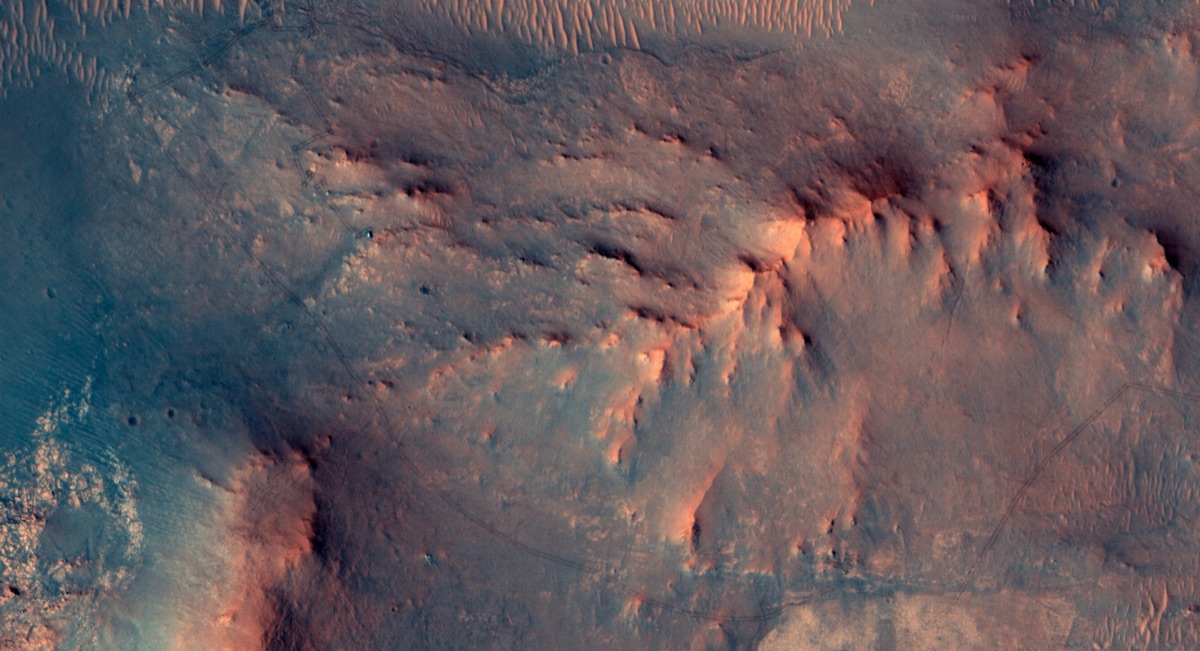 Overhead view from NASA's Mars Reconnaissance Orbiter (HiRISE camera) showing the Perseverance rover as a small bright speck on the rust-colored ground west of Jezero Crater, captured June 13, 2026, one day before the rover completed a marathon distance of driving on Mars.